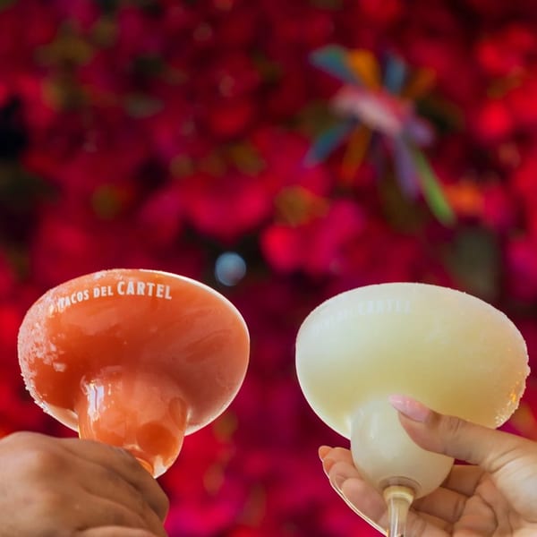 Two hands holding frozen Tacos Del Cartel margaritas, one orange and one yellow, against a red floral background.