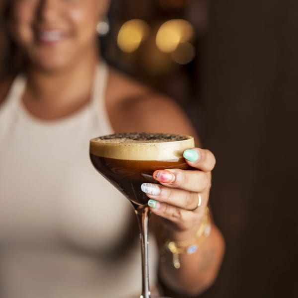 A smiling woman holds out a martini glass filled with a dark, foamy espresso martini.