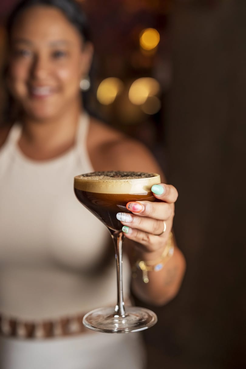 A smiling woman holds out a martini glass filled with a dark, foamy espresso martini.