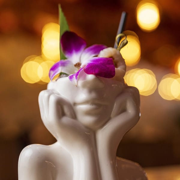 A cocktail in a white ceramic face-shaped mug, garnished with purple orchids and a shrimp skewer.