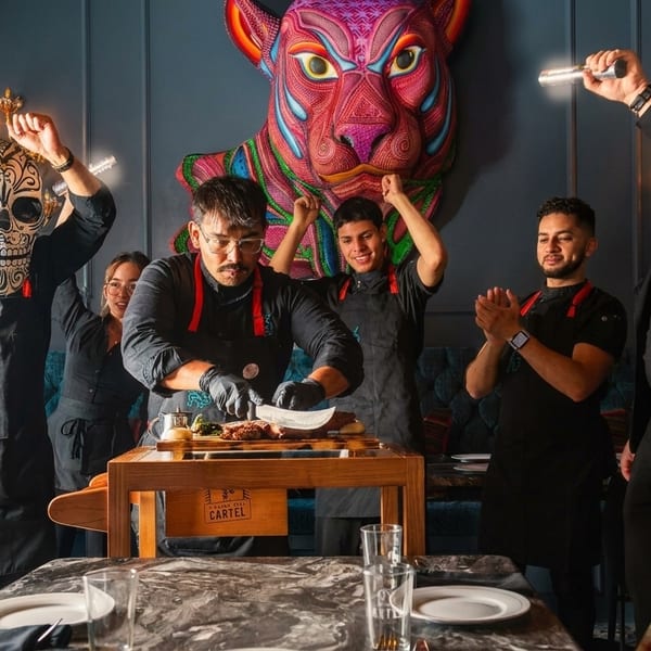 Restaurant staff celebrating a chef carving meat at a table, with a colorful jaguar art piece in the background.