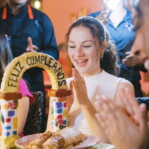 Smiling woman clapping at a table with a "Feliz Cumpleaños" arch and food.