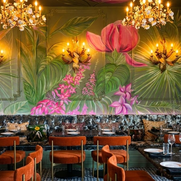 Restaurant interior with tropical mural, ornate chandeliers, black marble tables, and orange velvet chairs.