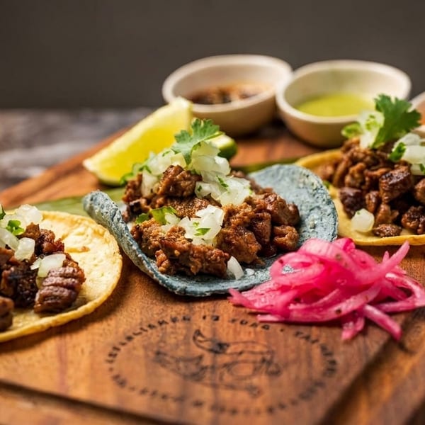 Three street tacos with meat, onions, cilantro, and lime on a wooden board with sauces.