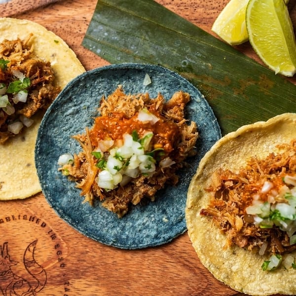 Three carnitas tacos with onions and cilantro on a wooden board with lime wedges and sauces.