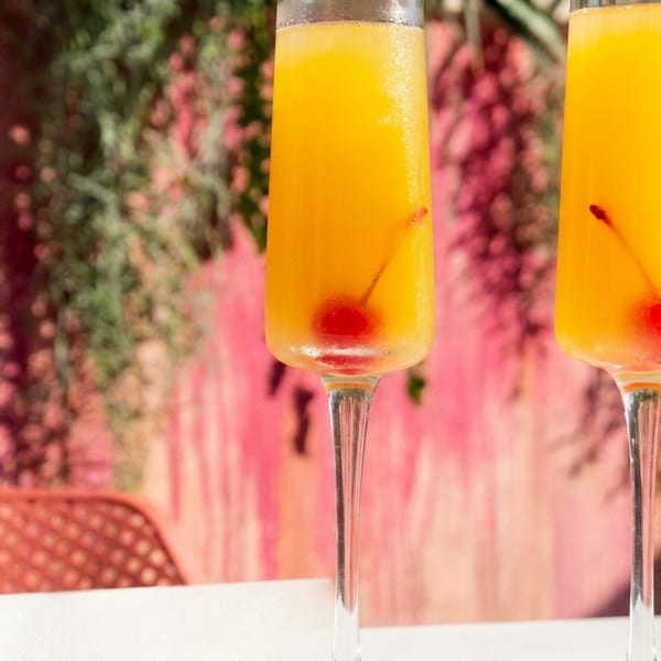 Two mimosa cocktails with cherries in champagne flutes on a white table with a pink background.