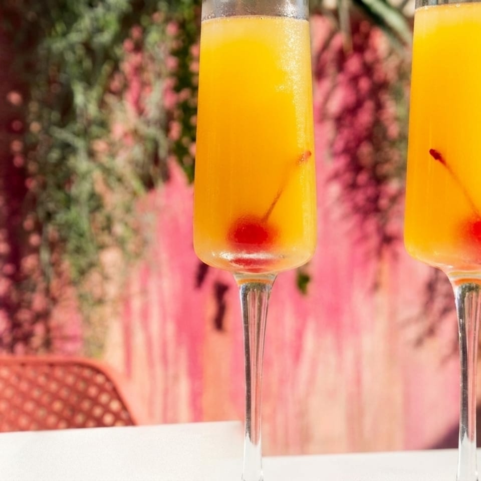 Two mimosa cocktails with cherries in champagne flutes on a white table with a pink background.