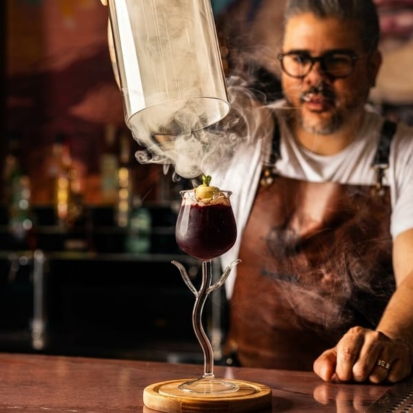 Bartender smoking a purple cocktail in a unique glass with a cloche, creating a dramatic effect.