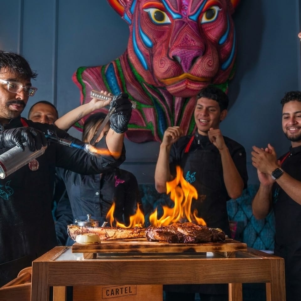 Chefs flambéing steaks at a table, with a colorful jaguar art piece in the background.