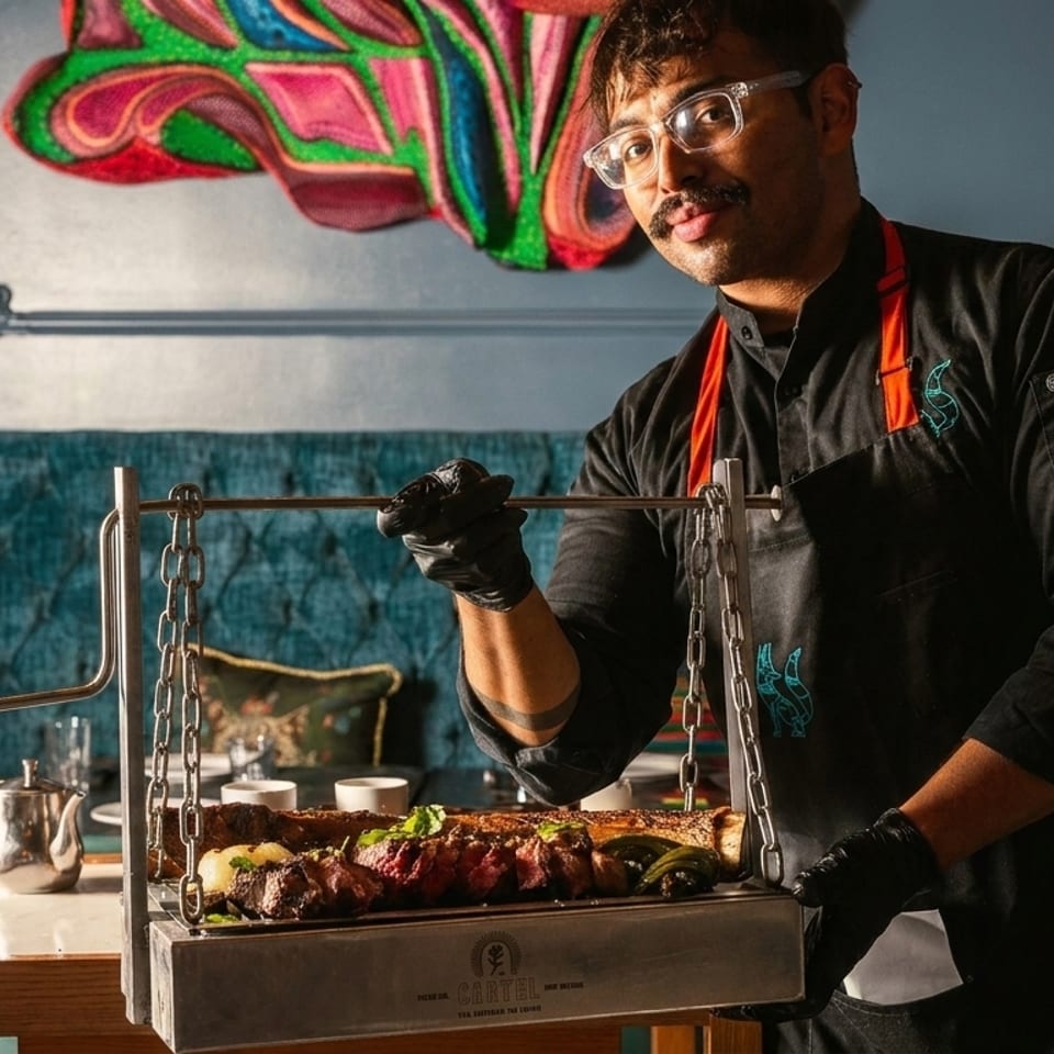 Chef in black uniform and glasses holding a hanging grill with cooked meat and vegetables.