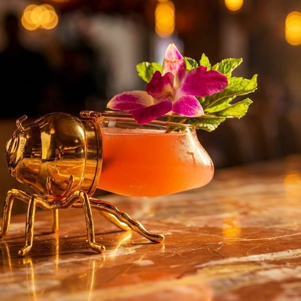 A vibrant orange cocktail in a golden beetle-shaped glass, garnished with a purple orchid and mint leaves.