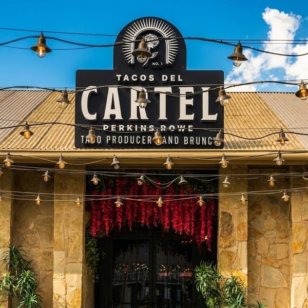 Tacos Del Cartel restaurant exterior with string lights, stone facade, and red floral decor.