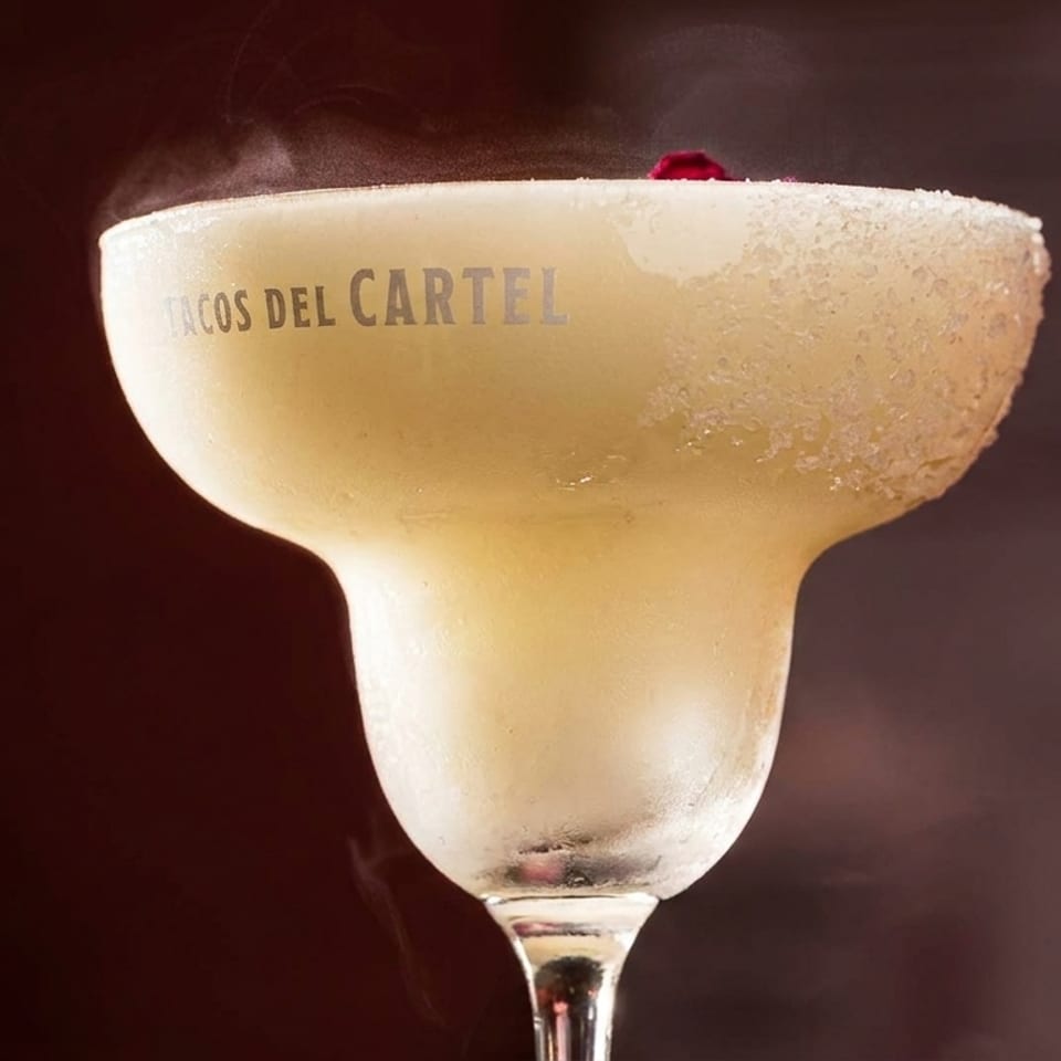 A frosty margarita in a branded glass with a salted rim and a red garnish, emitting vapor.